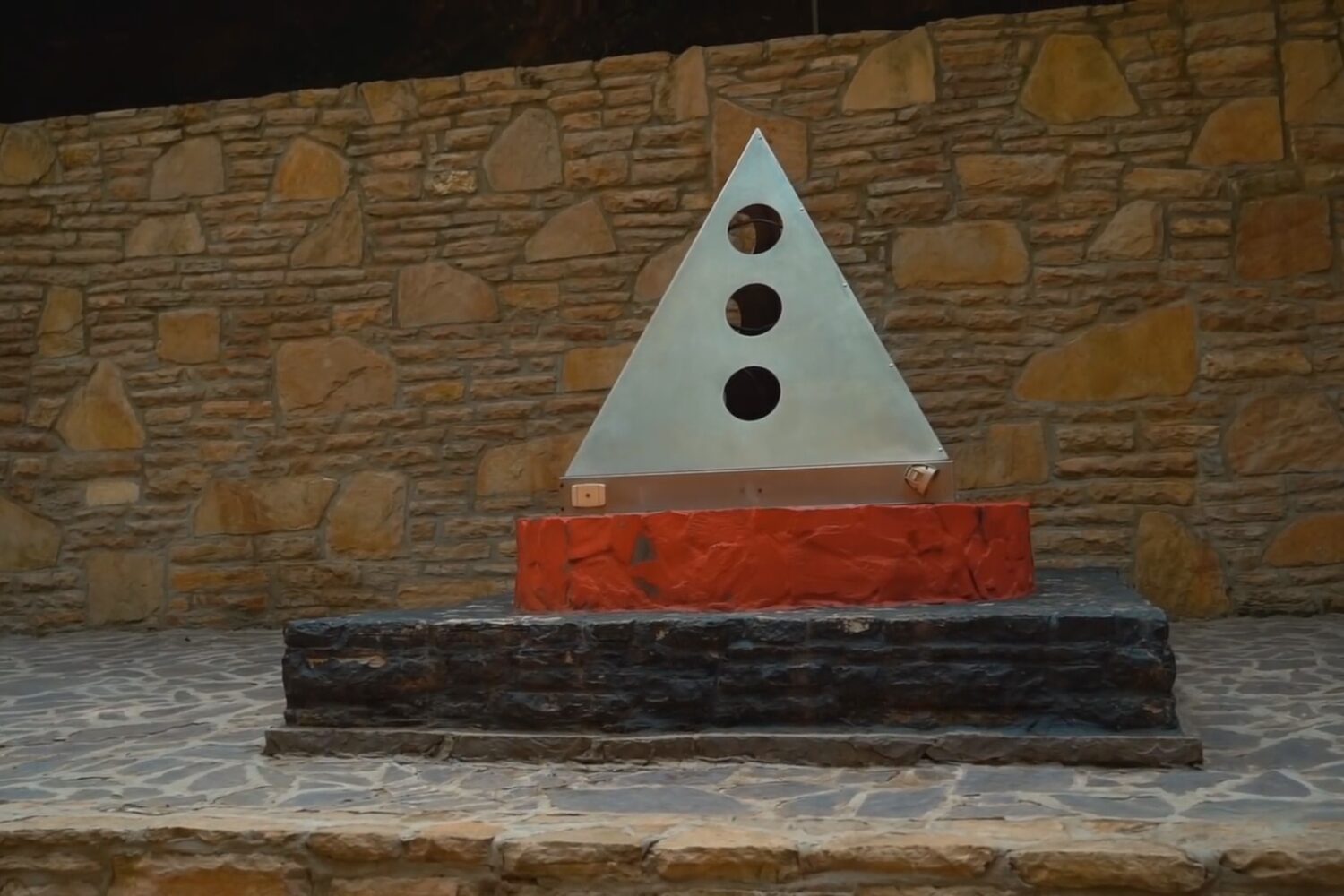 A metal pyramid sculpture with three circular cutouts, displayed on a tiered stone platform at the Bosnian Pyramid complex.