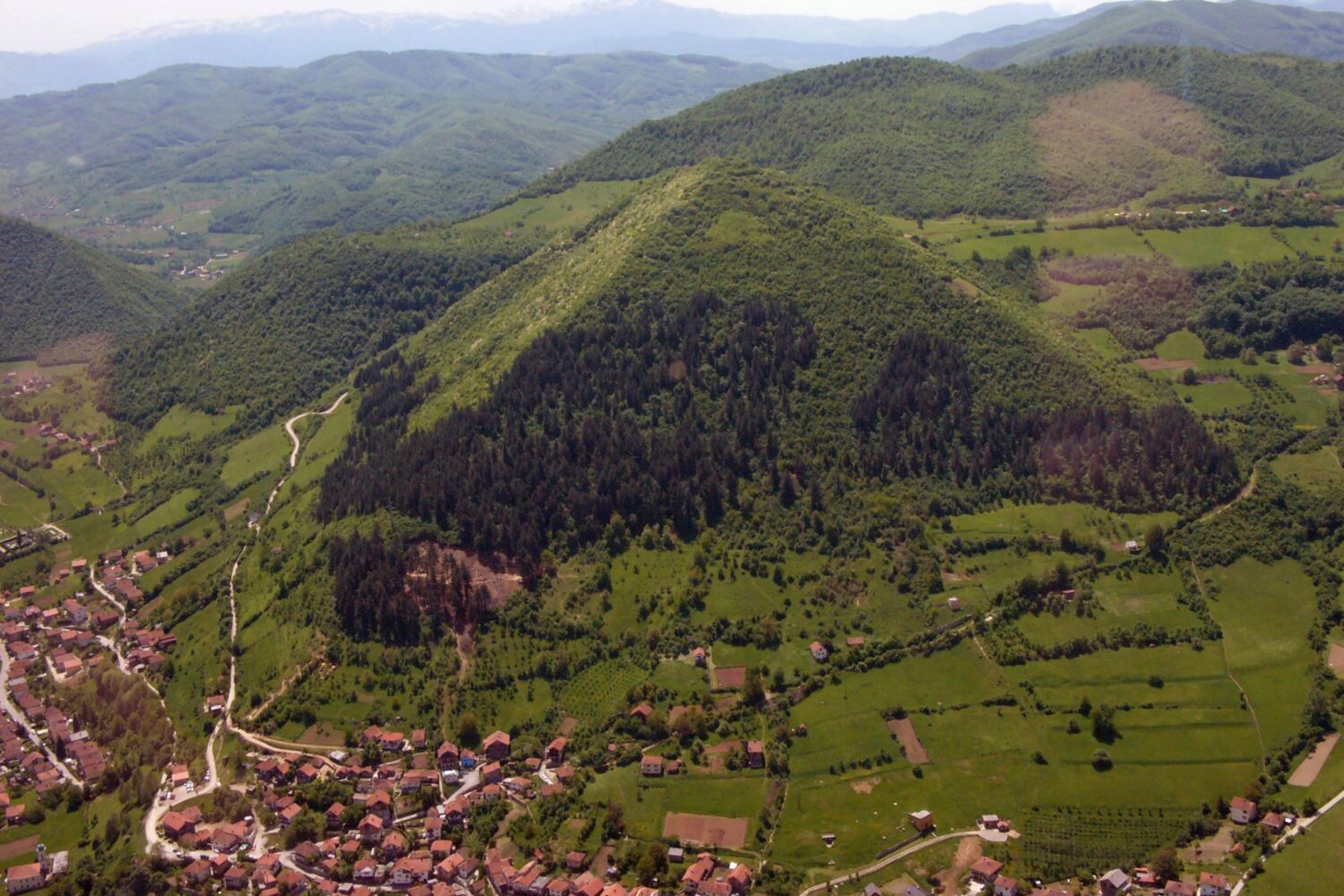 Aerial view of the Bosnian Pyramid of the Sun surrounded by lush greenery and a nearby village.