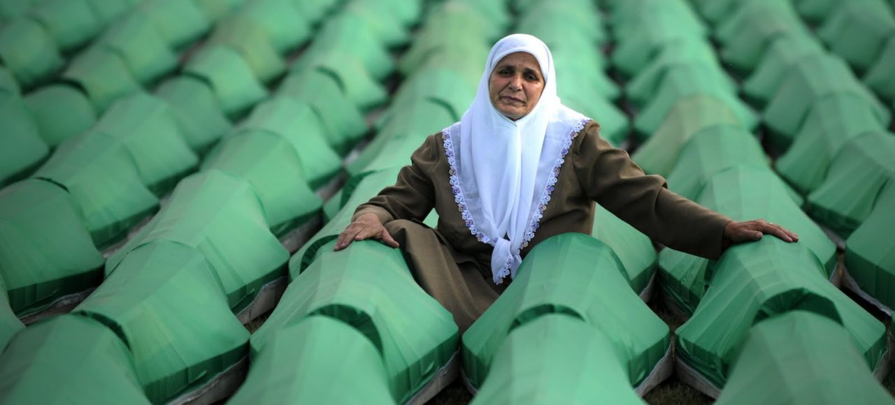 An elderly woman in a white headscarf sits among green coffins containing remains of Srebrenica massacre victims, awaiting burial.