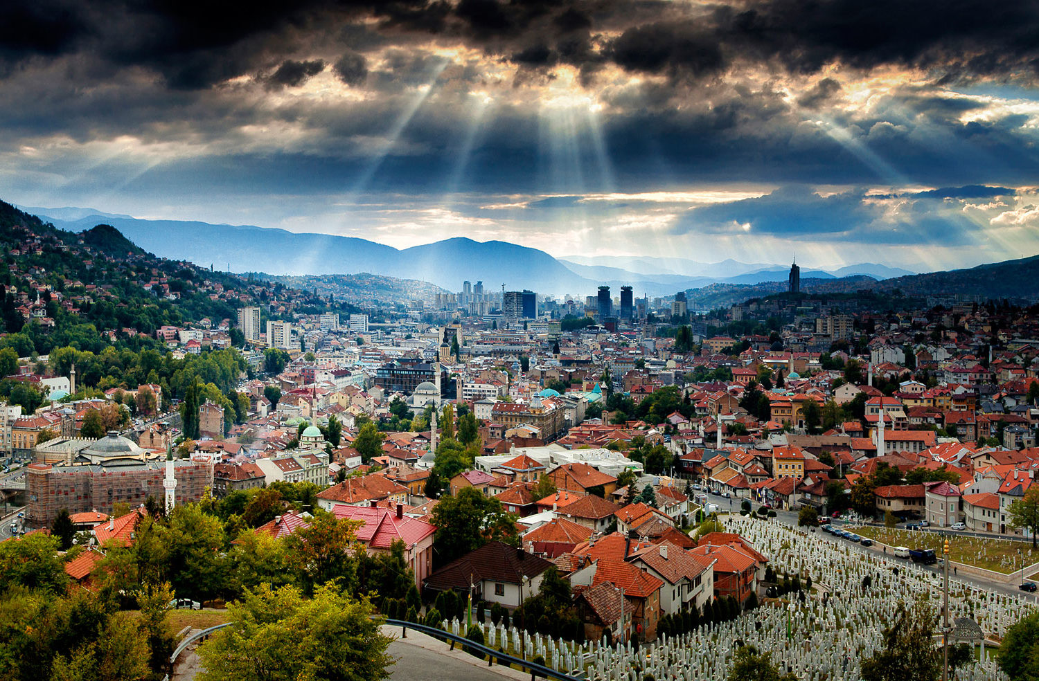 Panoramic view of Sarajevo city from a high vantage point, showcasing the city's dense architecture, surrounding mountains, and dramatic clouds with sun rays breaking through.