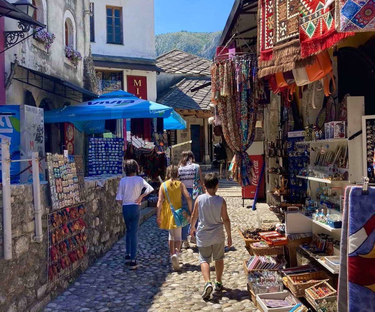 Children exploring a vibrant market street in Mostar, filled with colorful carpets and souvenirs.