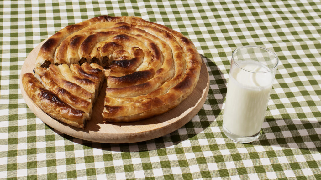 A freshly baked spiral-shaped burek on a wooden board, served with a glass of yogurt on a green-and-white checkered tablecloth.