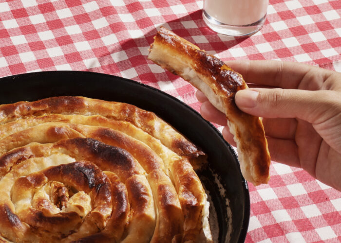 A freshly baked spiral-shaped burek in a cast iron pan, served with a glass of yogurt on a red-and-white checkered tablecloth.