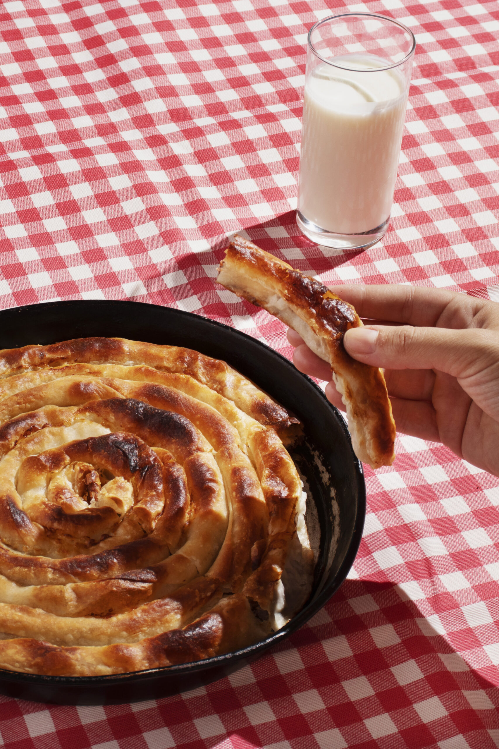 A freshly baked spiral-shaped burek in a cast iron pan, served with a glass of yogurt on a red-and-white checkered tablecloth.